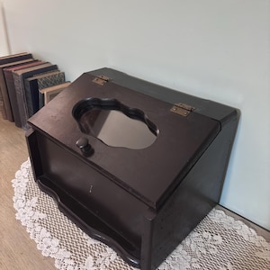 May include: A dark brown wooden bread box with a decorative cutout on the lid. The box has a hinged top and a small knob. The box is sitting on a white lace doily.