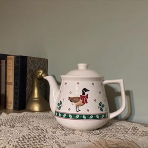 May include: White ceramic teapot with a lid, featuring a goose design with a red bow and holly accents. The teapot has a green and red border. A decorative item for the kitchen or dining room.