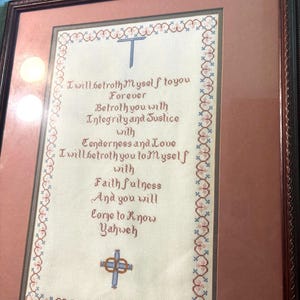 May include: Framed cross-stitch artwork with a dark brown frame. The artwork features a poem with a cross at the top and bottom, with text in shades of brown, blue, and pink. The text reads "I will betroth Myself to you Forever..."