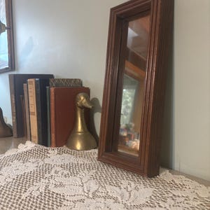 Puede incluir: Un espejo rectangular con marco de madera marrón se apoya sobre un mantel de encaje blanco. Detrás del espejo hay una figura de pato de latón y una pila de libros. El espejo refleja una ventana y un paisaje exterior.