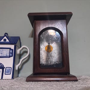 May include: A white and blue ceramic teapot shaped like a house, next to a dark wooden decorative box with a windmill scene. The windmill scene features a yellow sun and a black silhouette of a windmill.