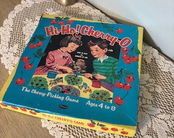 Puede incluir: Caja de juego de mesa vintage Hi-Ho! Cherry-O. La caja es azul con un borde amarillo y presenta ilustraciones de niños recogiendo cerezas. El juego es para niños de 4 a 8 años. El texto "Hi-Ho! Cherry-O" se muestra de forma destacada.