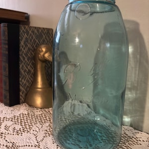 May include: A vintage blue-green Mason jar with embossed lettering. The jar is made of glass and has a wide mouth. The word "MASON" is clearly visible. The jar is sitting on a white lace tablecloth.