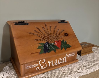 May include: A wooden bread box with a slanted lid and black metal hinges. The front features a hand-painted design of wheat stalks, purple grapes, and a red apple, with the word Bread written in white cursive script across the bottom.