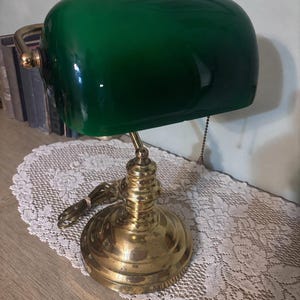 May include: A banker's lamp featuring a polished brass base and a curved, emerald green glass shade. The lamp includes a pull chain switch hanging from the side of the shade and a brass arm that connects the shade to the circular, tiered base.