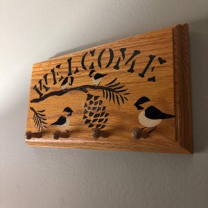 Puede incluir: Letrero de bienvenida de madera con letras negras, una piña y ilustraciones de pájaros. El letrero tiene tres clavijas de madera para colgar objetos. El letrero es de madera de color marrón claro.