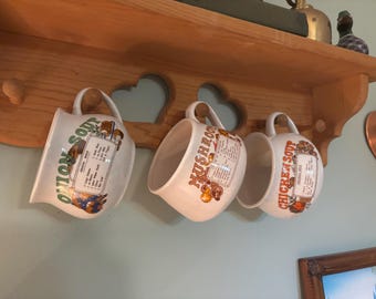 Puede incluir: Tres tazas de sopa de cerámica blanca colgadas de un estante de madera. Cada taza presenta una etiqueta de sopa diferente: Sopa de cebolla, Sopa de champiñones y Sopa de pollo. Las tazas tienen un diseño clásico y vintage.