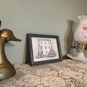 Puede incluir: Un sujetalibros con forma de pato de latón se encuentra junto a un cuadro enmarcado con una ilustración de gato y una lámpara de aceite blanca con rosas rosas. La lámpara se encuentra sobre una base blanca y tiene una parte superior con volantes. Los artículos están sobre un mantel de encaje.