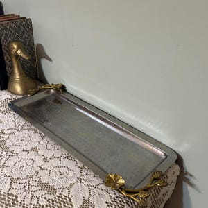 Puede incluir: Una bandeja rectangular plateada con asas florales doradas. La bandeja tiene una superficie texturizada y se exhibe sobre un mantel de encaje blanco. Una figura de pato dorado es visible en el fondo.