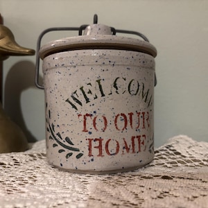 Puede incluir: Un tarro de cerámica moteada con tapa y asa metálica. El tarro está inscrito con las palabras "WELCOME TO OUR HOME" en letras verdes y rojas. A la izquierda se ve una figura de pato de latón.