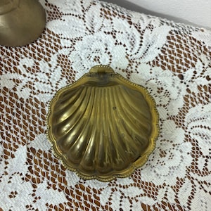 May include: A brass trinket box shaped like a scallop shell. The container features a hinged lid with a ribbed, fan-like texture and a decorative beaded edge. The metal surface has a polished, golden finish with subtle signs of natural aging.