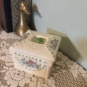 May include: A cream-colored, rectangular trinket box with a lid. The box features a ribbed design and is adorned with floral patterns in pink, blue, and green. A green, round knob sits atop the lid. The box is on a lace tablecloth.