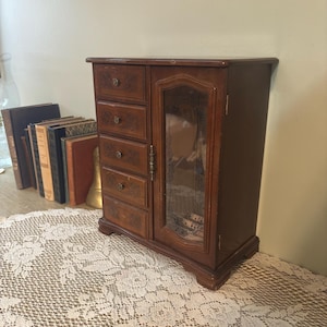 Puede incluir: Un joyero de madera con acabado marrón oscuro. Cuenta con cuatro cajones pequeños en el lado izquierdo con tallas decorativas y una puerta con frente de vidrio en el lado derecho. El joyero está sobre un mantel individual de encaje, con libros visibles en el fondo.