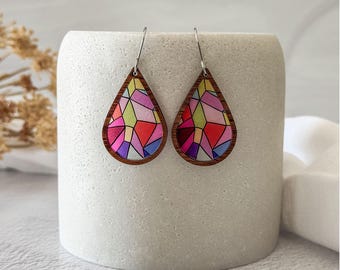 May include: A pair of teardrop-shaped earrings with a stained-glass design. The earrings feature a wooden frame and a colorful mosaic pattern in shades of pink, purple, yellow, and red. The earrings are displayed on a white cylindrical object.