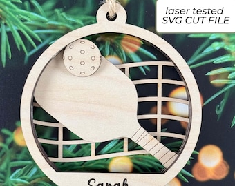 May include: A circular wooden ornament featuring a cutout design of a pickleball paddle, a ball, and a net. The name Sarah is engraved at the bottom. A jute twine loop is attached to the top. The text laser tested SVG CUT FILE is visible.