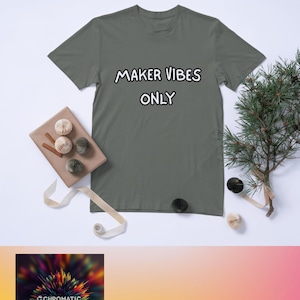 以下が含まれることがあります： 手書き風の「MAKER VIBES ONLY」の文字が入ったグレーのTシャツ。白い表面に、紙のオーナメントや小さな常緑樹の枝など、装飾品と一緒に展示されています。