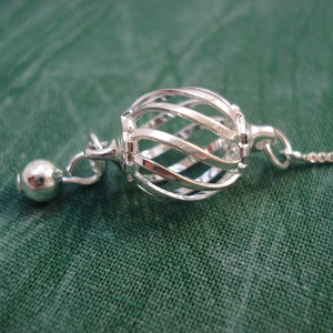 May include: A silver chain necklace with a silver cage pendant. The cage is made of twisted wire and has a small silver ball at the end.
