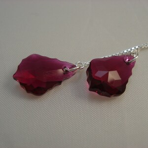 May include: A necklace featuring two faceted, irregular-shaped, deep magenta crystal pendants. The pendants are connected to a delicate silver chain. The crystals have a sparkling, jewel-like appearance.