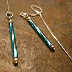 May include: A pair of dangle earrings featuring teal blue, cylindrical beads with silver-tone accents. The earrings have a delicate chain design, with one earring having a long, straight post. The jewelry is displayed on a textured, dark surface.