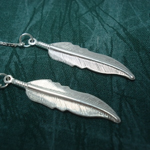 May include: A pair of silver feather earrings with delicate chains. The feathers are detailed with fine lines and have a slightly textured surface.