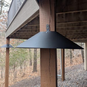 May include: A matte black metal light fixture cover with a wide, conical shade design. The fixture is mounted directly onto a vertical wooden post using a small metal bracket secured with two visible screws at the top center of the shade.