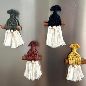 May include: Five handmade macrame gnome ornaments with cinnamon stick beards. Each gnome has a different colored hat: green, black, burgundy, and yellow. The gnomes have white beards and small wooden bead noses. They are displayed on a silver surface.