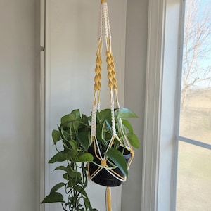 Puede incluir: Un colgador de plantas de macramé blanco y amarillo, sosteniendo una maceta negra llena de plantas verdes. El diseño de macramé presenta nudos intrincados y una borla, suspendido cerca de una ventana.