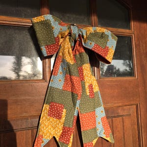 Könnte beinhalten: Eine dekorative Stoffschleife mit Patchwork-Design in verschiedenen Mustern in den Farben Olivgrün, Rostorange, Hellblau und Gelb. Die Schleife hat zwei lange, spitz zulaufende Enden und besteht aus gestepptem Stoff mit weißen Punkten.