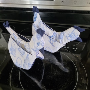 Peut inclure: Deux maniques en forme de poulet, blanches et bleues. Les maniques présentent un motif bleu et blanc de poulets et de paniers. Elles ont une crête et un bec bleu foncé. Les maniques sont posées sur une plaque de cuisson noire.