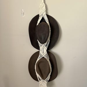 Puede incluir: Un perchero para sombreros de macramé blanco con dos sombreros de vaquero marrones colgados. El perchero está sujeto a una pared blanca con un gancho de plástico transparente.