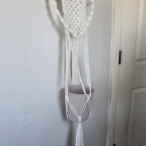 May include: A white macrame plant hanger with a beige striped pot. The hanger is suspended from a wooden dowel and features intricate knotting and a tassel. The pot is designed to hold a plant.
