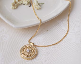 Può includere: Una collana color oro con un pendente circolare. Il pendente presenta una pietra centrale trasparente, circondata da un motivo floreale e un bordo di pietre trasparenti più piccole. La collana è su un piatto bianco con fiori secchi.