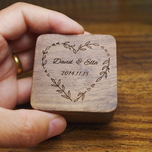 May include: A square wooden ring box featuring a heart-shaped wreath design. The box is engraved with the names "David & Ella" and the date "2014.11.15". The box is held in a hand, and the wood has a natural grain.