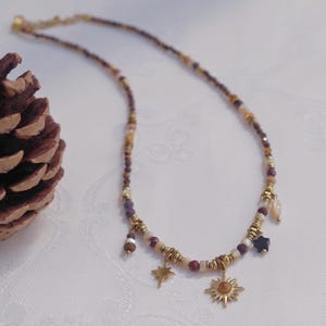 May include: A beaded necklace featuring small multicolored stones in shades of brown, cream, and purple, accented with gold-toned metal spacers. The necklace includes dangling charms shaped like a star, a sun with a center stone, and a dark star.