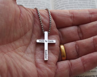 Puede incluir: Un collar con colgante de cruz plateada con las palabras "Walk by Faith" y "Ezekiel" grabadas. La cruz cuelga de una cadena plateada. El fondo incluye una mano y un libro abierto.