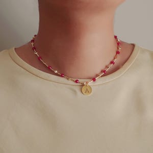 Può includere: Collana dorata con una catena delicata, con piccole perline rosse e un ciondolo rotondo. Il ciondolo ha incisa la lettera "A". La collana è indossata intorno al collo.