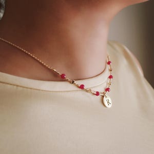 Könnte beinhalten: Goldfarbene Halskette mit zarter Kette und kleinen roten Perlen. Die Halskette hat einen Anhänger mit dem eingravierten Buchstaben "R". Die Halskette wird um den Hals getragen.