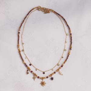 May include: A layered necklace set featuring one strand of small, multi-colored earth-toned beads with gold-tone accents and charms including a sun, a star, and a small stone, paired with a delicate gold-tone chain featuring small, flat circular discs.