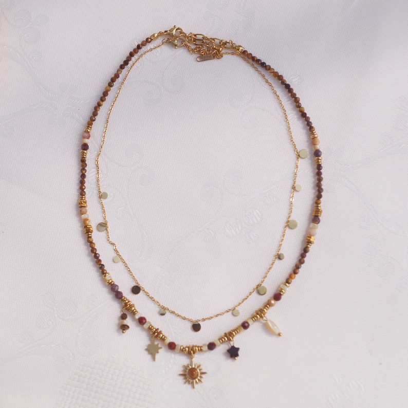 May include: A layered necklace set featuring one strand of small, multi-colored earth-toned beads with gold-tone accents and charms including a sun, a star, and a small stone, paired with a delicate gold-tone chain featuring small, flat circular discs.