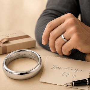 May include: A polished silver-toned metal ring featuring the engraved text "Here with you. E.J" followed by a heart symbol. The same inscription is written on a piece of paper nearby, and a similar ring is worn on a finger in the background.
