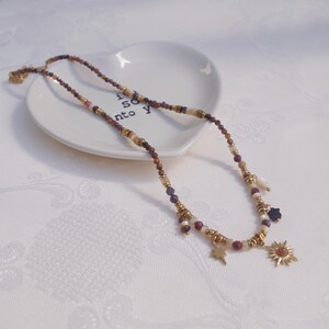 May include: A beaded necklace featuring small, multi-colored faceted stones in shades of brown, purple, and cream, interspersed with gold-toned metal spacers. The necklace includes dangling charms shaped like a star, a sun, and a small black star.