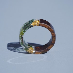 Könnte beinhalten: Ein handgefertigter Ring mit einem einzigartigen Design. Die Hälfte des Rings besteht aus dunklem Holz, während die andere Hälfte aus klarem Harz mit grünen Pflanzeneinschlüssen und Goldblattakzenten besteht. Der Ring hat eine kreisförmige Form.