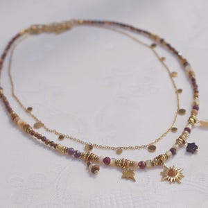 May include: A layered necklace featuring a delicate gold chain with small circular discs and a second strand composed of multi-colored beads in shades of brown, cream, and burgundy, accented with gold spacers and charms shaped like a star and a sun.