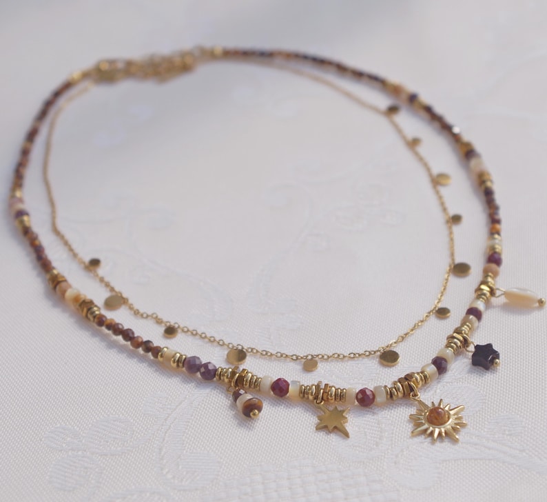 May include: A layered necklace featuring a delicate gold chain with small circular discs and a second strand composed of multi-colored beads in shades of brown, cream, and burgundy, accented with gold spacers and charms shaped like a star and a sun.