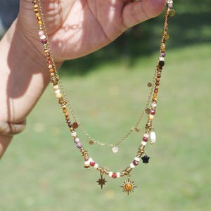 May include: A layered necklace featuring two gold-toned chains. The longer chain is adorned with a variety of small, multi-colored beads in shades of brown, white, and red, interspersed with gold-toned spacers, a black star charm, and sun charms.