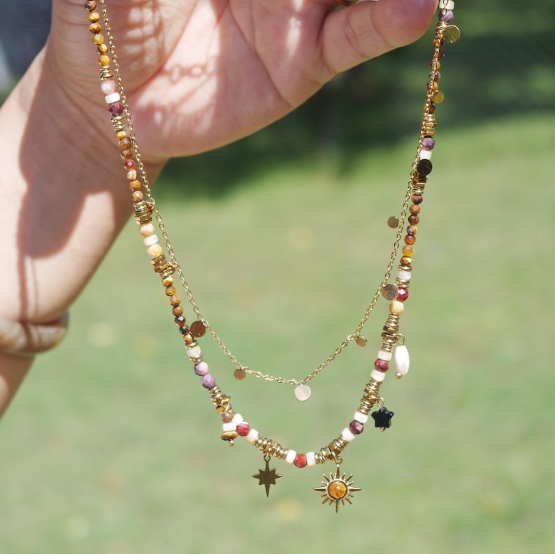 May include: A layered necklace featuring two gold-toned chains. The longer chain is adorned with a variety of small, multi-colored beads in shades of brown, white, and red, interspersed with gold-toned spacers, a black star charm, and sun charms.