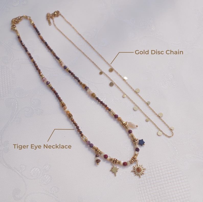 May include: A Tiger Eye necklace featuring multi-colored beads, gold-toned spacers, and charms including a star and a sun, alongside a gold disc chain necklace. The image includes the text labels Tiger Eye Necklace and Gold Disc Chain.