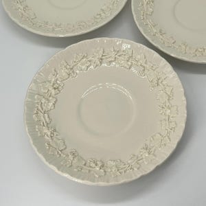 May include: Three off-white ceramic saucers with a raised floral design around the rim. The saucers are arranged on a white surface, each with a central indentation for a cup. The floral pattern adds a touch of elegance.
