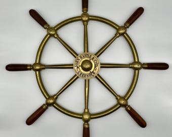 May include: A brass ship's wheel with eight spokes and dark brown wooden handles. The centre of the wheel features a circular emblem with the text "JOHN HASTIE & CO. GREENOCK". This nautical decorative item is a ship's wheel.
