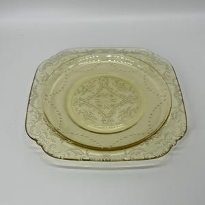 May include: A set of two yellow-tinted glass plates. The larger plate is square with a scalloped edge, and the smaller plate is round. Both plates feature an embossed floral and geometric design.